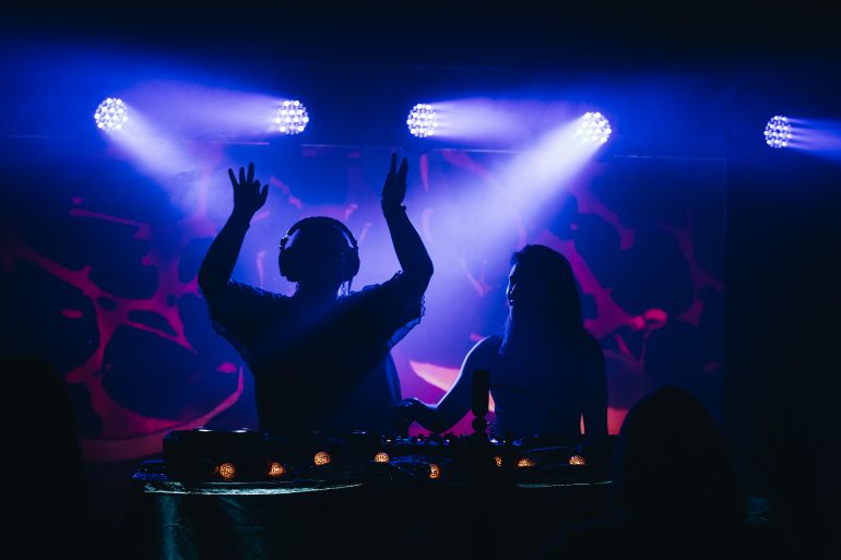 Two figures in shadow DJing on a stage one with her arms up.