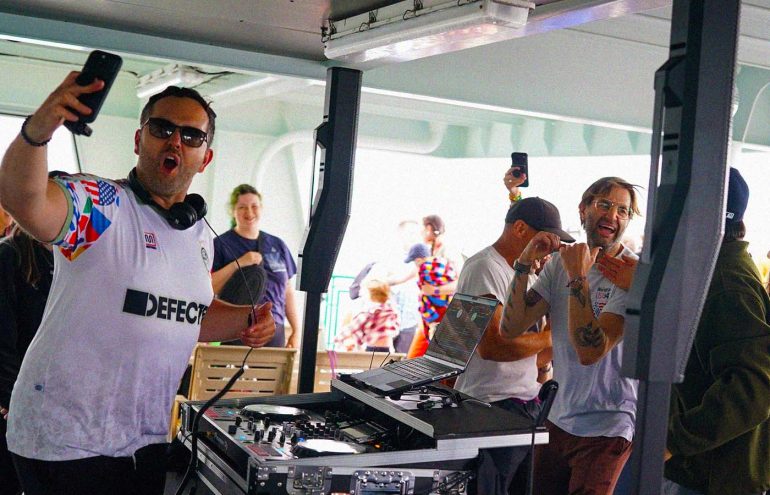 A person wearing a "Defected" shirt takes a selfie near DJ equipment on a deck. Another person in the background appears excited, holding a drink. Several others are seen smiling and mingling.