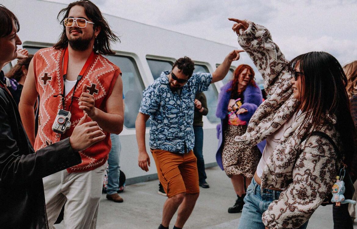 A group of people dancing energetically on a boat deck. One person wears a red vest with patterns, another in a blue floral shirt dances in the center. A person in leopard print and colorful attire holds a plush toy. The background shows the boat's structure and a cloudy sky.